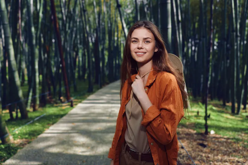 Woman with backpack smiling on a forest path, portrait of a young hiker outdoors surrounded by trees and sunlight, casual clothing and relaxed travel mood in nature.