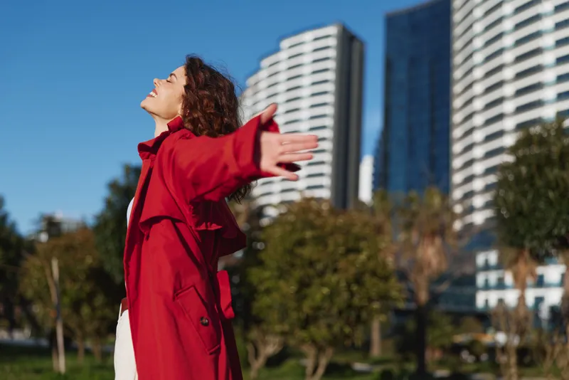 A woman in a red coat stands outdoors with arms outstretched, enjoying sunny city life. Bright buildings frame a moment of freedom and joy.