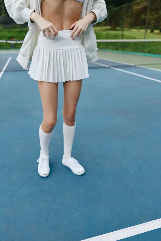 Young athletic woman wearing a white skirt and sports socks on a tennis court, showcasing confidence and energy in an outdoor sports setting