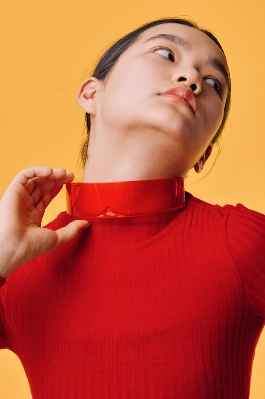 Woman in red sweater and matching shiny choker posing with head turned on orange background for fashion portrait and style concept in studio lighting.