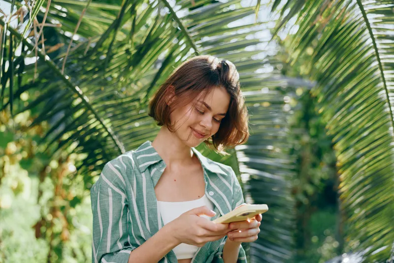 Young woman with short brown hair stands outdoors under palm fronds, smiling softly while looking down at a handheld device.