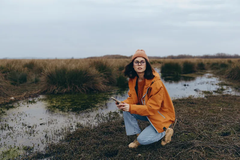 Woman lifestyle outdoors portrait, kneeling by river water and grass, orange jacket, knit hat and glasses, looking at camera in an autumn countryside field near muddy shore.