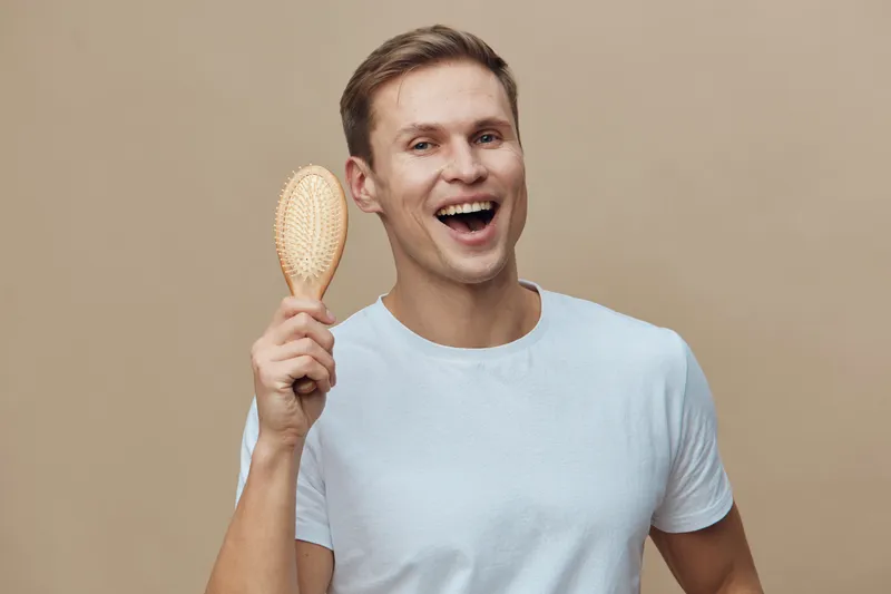 Happy smiling man with short brown hair wearing a light blue t-shirt holding a hairbrush near his head, standing on beige background in a studio portrait. People lifestyle concept, grooming, self-care