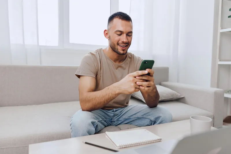 Engaged man using smartphone, sitting comfortably on a sofa in a bright, modern living room, showcasing a productive workspace with a notebook and coffee.