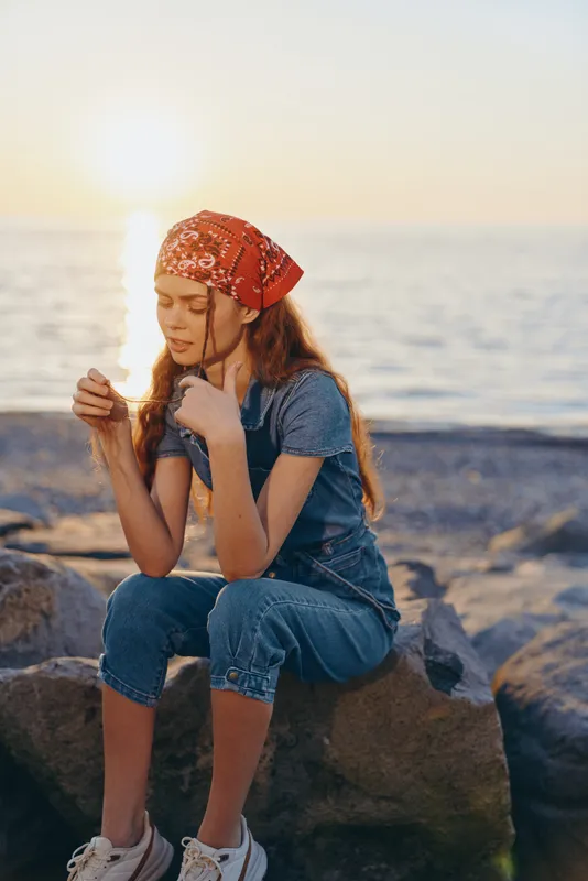Lifestyle woman in modern boho-western denim street style with red bandana, sitting on rocks by the sea during golden hour, film color tones emphasizing warm natural light.