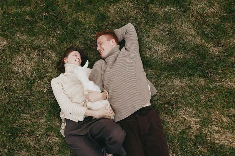 Couple grass outdoors dog lifestyle affection companionship relaxed moments natural. A candid outdoor scene shows a smiling pair lying on grass with a small dog, sharing warmth and playful tenderness