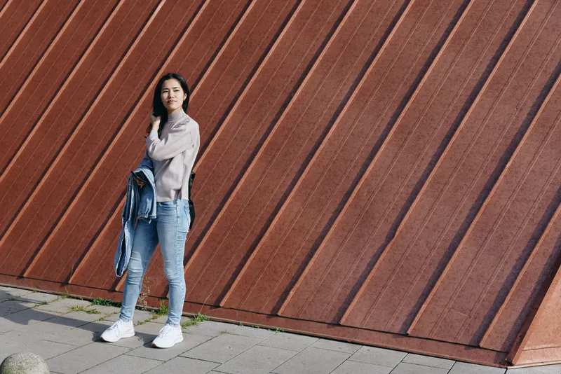 Woman standing against a diagonal textured wall in casual street style, wearing jeans, sneakers and a light jacket, holding a denim jacket for a city stroll
