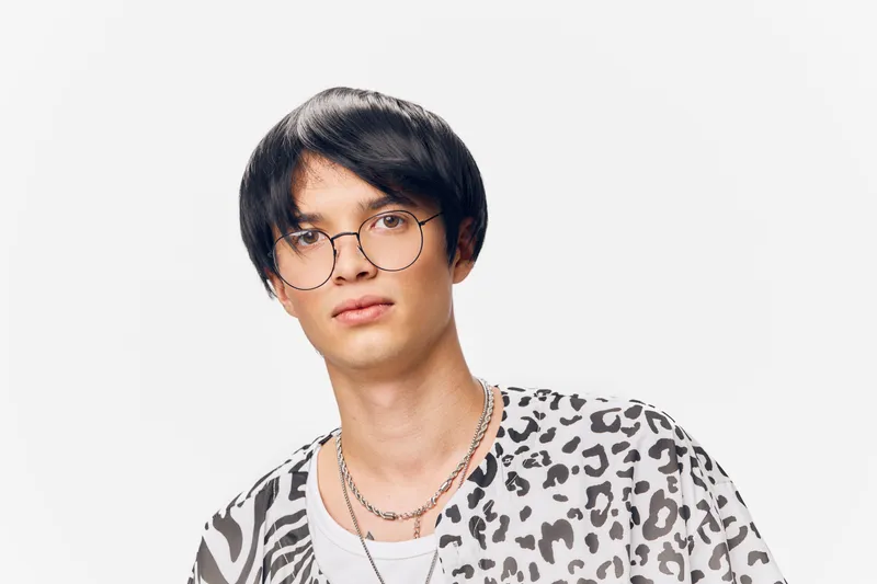 young man wearing round glasses and animal print shirt with layered necklaces on white background, casual style and natural expression