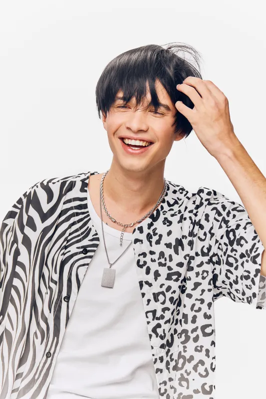 Smiling man with black hair wearing a patterned shirt and white t-shirt, showing joyful expression against a bright white background in a casual style.