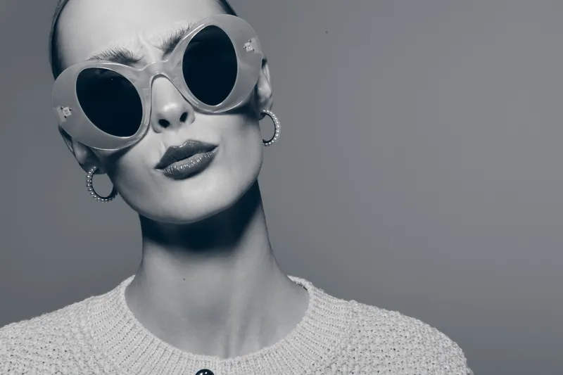 Stylish caucasian woman with pale skin wearing round sunglasses and earrings looks confidently in studio.