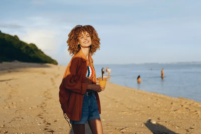 Woman beach ocean sunny summer travel lifestyle holding drink curly hair smiling, standing outdoors near shoreline, warm sand and calm sea, travel vacation mood, soft golden light