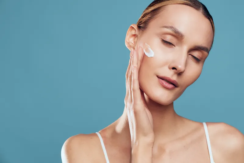 Young woman applying moisturizer on face with closed eyes, smooth skin texture, gentle touch, serene expression, blue background, skincare routine, natural beauty concept