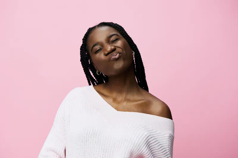 Happy young woman with braids in a stylish sweater against a pink background, showcasing confidence and positivity Perfect for lifestyle and fashion themes