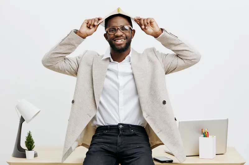 Smiling man wearing glasses and a light gray blazer holding a small cardboard box on his head, standing in a modern office with white background, cheerful mood, casual style, business casual, professi