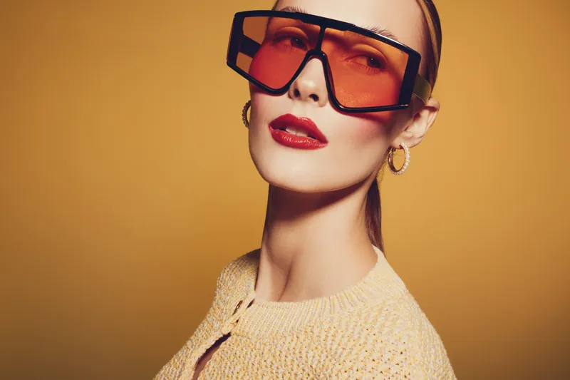 Stylish woman with oversized orange sunglasses and red lipstick poses against yellow background. Fashion portrait with trendy accessories and confident expression. Concept of modern style.