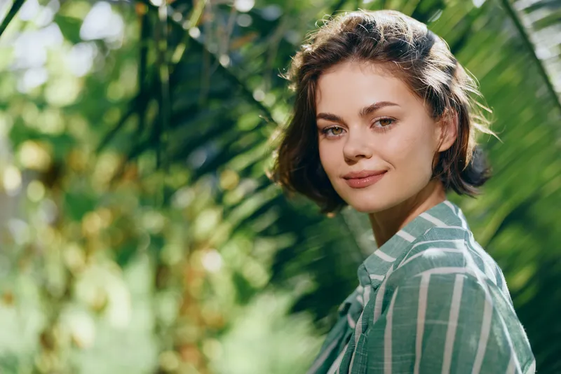 Portrait of a young woman with short brown hair in a lush tropical garden, soft sunlight, gentle smile, casual striped shirt, serene mood.
