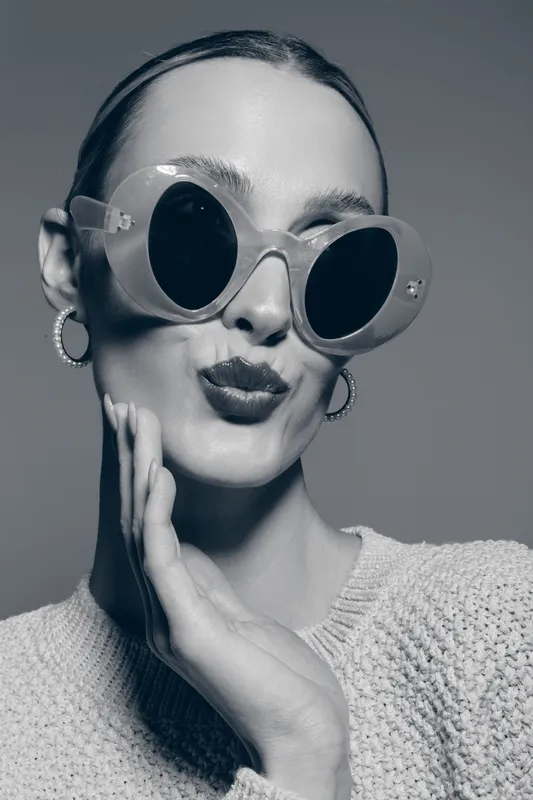 Stylish Caucasian woman with fair skin wearing large round sunglasses and hoop earrings poses with playful expression on neutral background. Fashion and portrait concept.