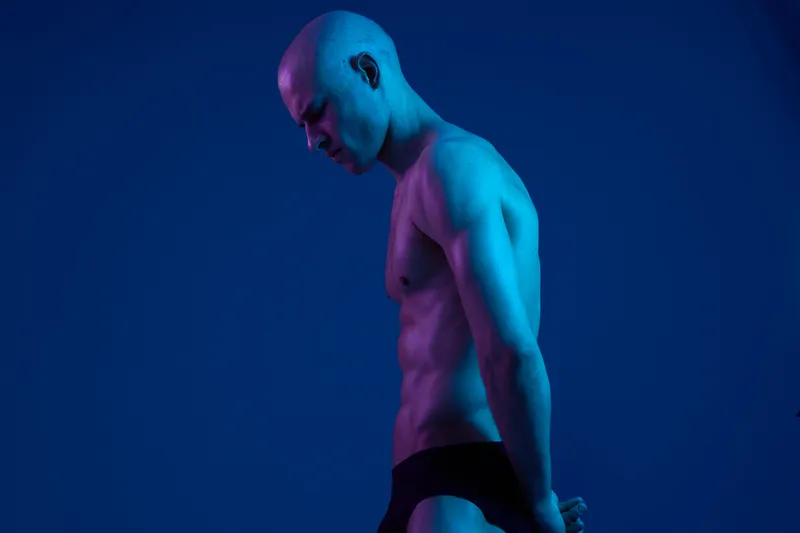A strong, muscular man with a shaved head poses in a blue-lit setting, highlighting his physique. This striking photo captures confidence and strength, perfect for fitness themes.
