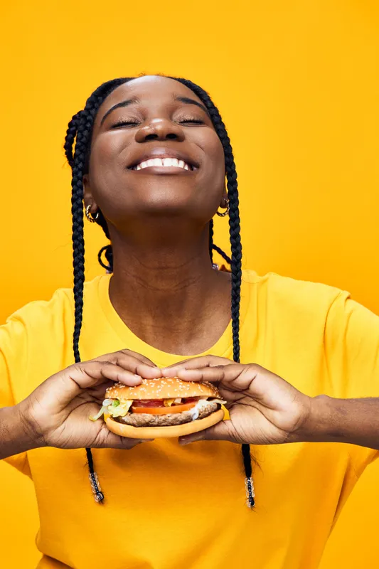 Happy young woman with braids enjoying a burger against a bright yellow background, symbolizing joy and indulgence with vibrant colors