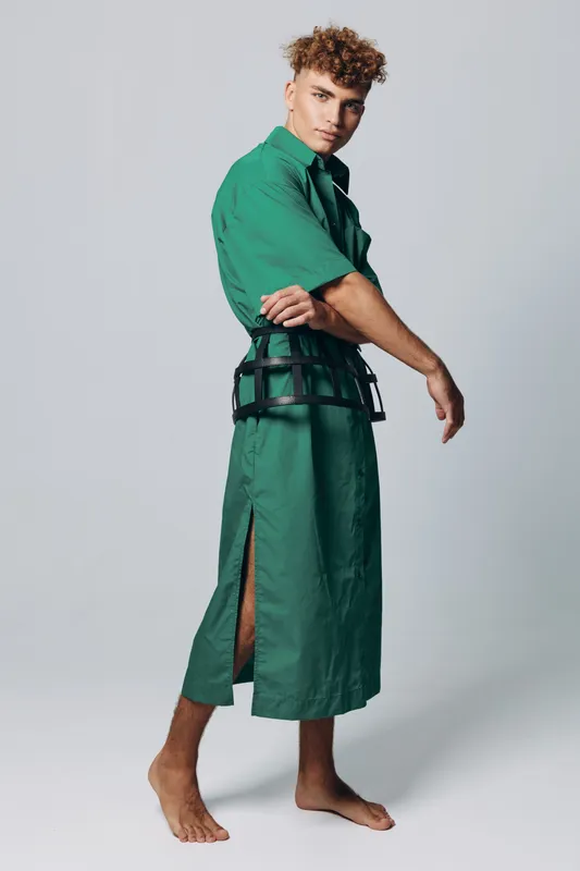 Young man in green long dress with side slit and black belt posing barefoot on gray background in studio. Fashion, style and casual outfit with curly hair and confident look.