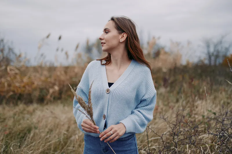A candid portrait of a woman in a cozy blue cardigan standing in a windswept meadow, conveying authenticity and quiet confidence through natural light and simple, truthful composition.