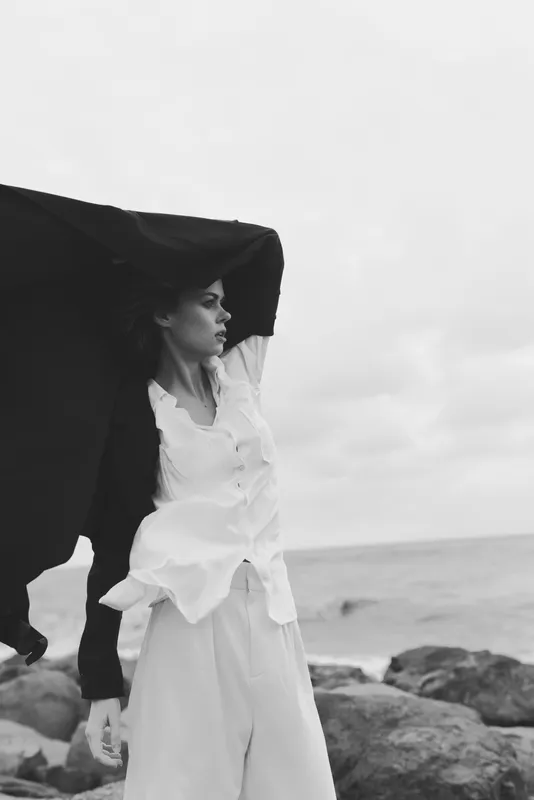 Woman in white dress standing on rocky shore with flowing cape in wind, black and white fashion shot capturing movement and ocean mood and dramatic lighting