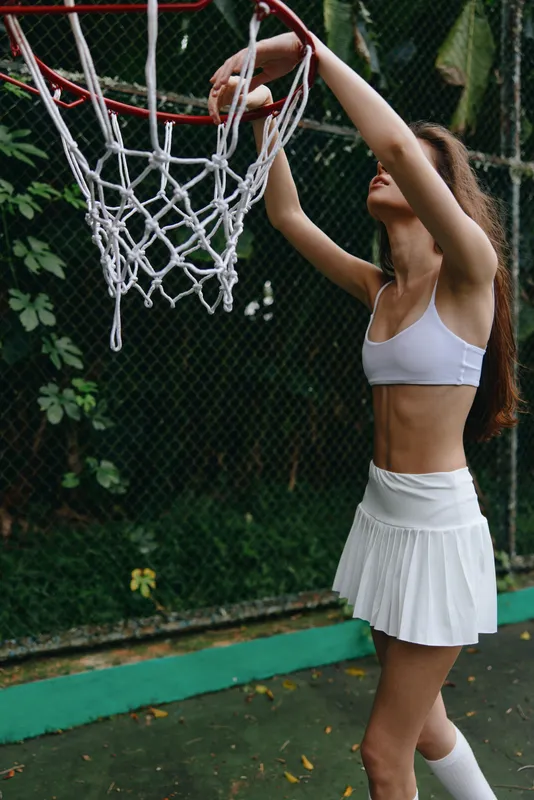 Sporty female athlete with a toned physique playing basketball outdoors in a green setting with stylish white sportswear, capturing determination and athleticism