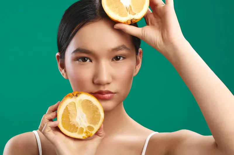 cheerful asian woman holds citrus halves against a vivid green backdrop, smiling with a lively expression that conveys freshness, energy, and a modern, vibrant vibe.