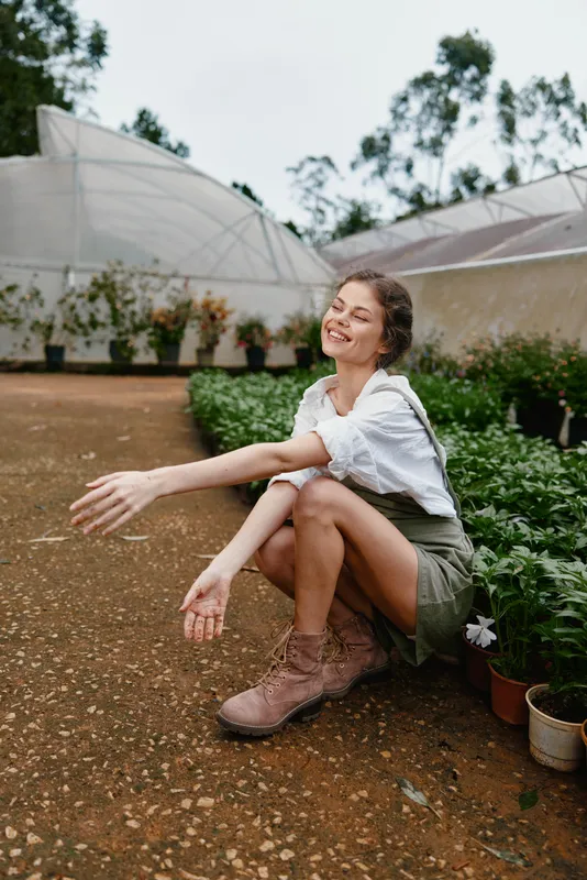 Happy young woman in stylish outfit sitting in greenhouse, enjoying nature, surrounded by plants, wearing boots, showcasing spring vibes and joy