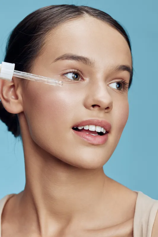 Beautiful young woman applying skincare serum for a healthy glow with a smile, showcasing natural beauty against a soft blue background