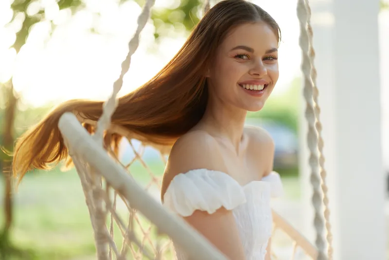 Young woman with long hair smiling on a swing in a sunny outdoor setting, wearing a white dress, conveying joy and summer vibes