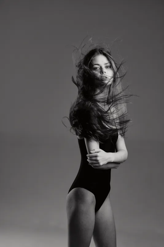 Black and white portrait of a young woman with flowing hair wearing a black bodysuit, expressing calm confidence in a studio setting.