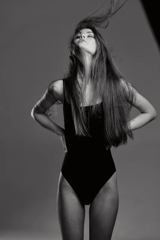 Black and white portrait of a slender woman with long hair in a black swimsuit posing confidently in a studio setting.