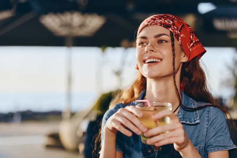 Lifestyle woman in modern boho-western denim street style smiling and enjoying a drink outdoors with vibrant film color tones and natural sunlight on a warm day.