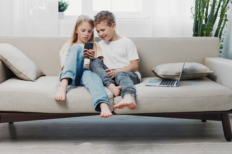 Two children sitting barefoot on a sofa, looking at a smartphone together in a bright living room. Concept of family, technology, childhood, and leisure time at home.