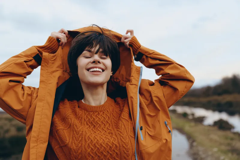 Woman lifestyle outdoors, young laughing windy moment, wearing jacket and sweater, hands on head, brunette hair moving, standing on coastal hill with ocean waves and clouds, travel joy