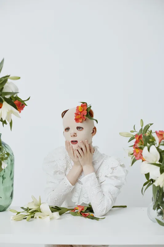 A woman wearing a facial mask with flowers, expressing surprise, set against a minimalist white background, featuring bright florals and a modern aesthetic