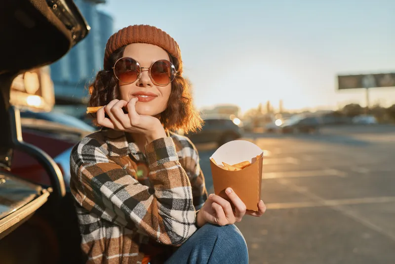 woman with fries by car at sunset wearing sunglasses and beanie in parking, smile and relaxed vibe capturing authenticity, candid lifestyle, golden hour glow, mindful living, emotional storytelling