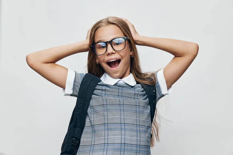 Girl with glasses and backpack smiling with open mouth in studio. Child wearing checkered dress and white shirt posing with hands on head for school portrait.