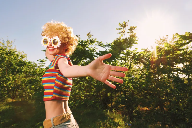 Playful woman with curly wig and flower sunglasses reaches toward the camera in a sunlit park, wearing a striped top, embodying carefree retro lifestyle and vibrant outdoors