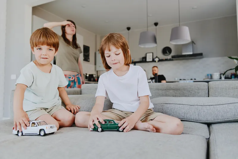 Playful children focused on toy cars in a cozy living room, while their mother watches from the background, creating a warm family atmosphere.