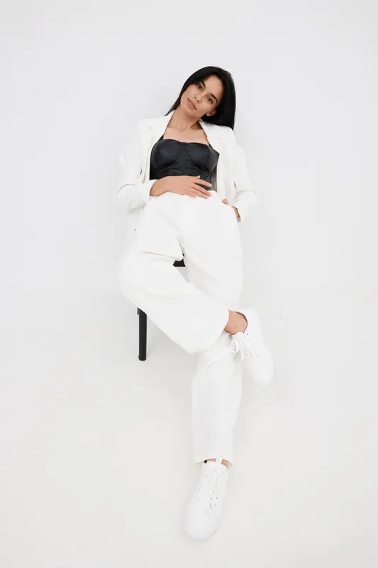 fashion woman wearing white suit and black top sits relaxed against white wall with casual pose showcasing modern style and confident attitude in minimalistic studio setting