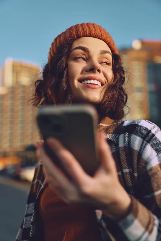 Woman smile with phone in urban street wearing beanie, candid moment bathed in golden hour glow, authenticity and mindful living detail for emotional storytelling lifestyle image.