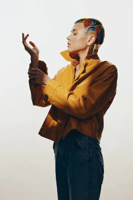 Woman with colorful shaved hairstyle wearing yellow jacket and blue jeans stands in profile striking a graceful pose against neutral background.