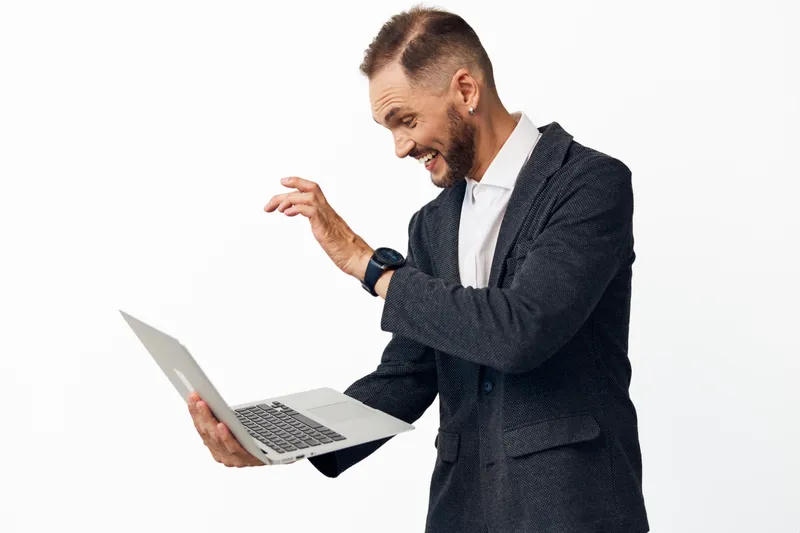 A professional man in a dark blazer stands against a clean white background, expressing excitement and positive energy as he peers at a laptop screen during a casual work moment.