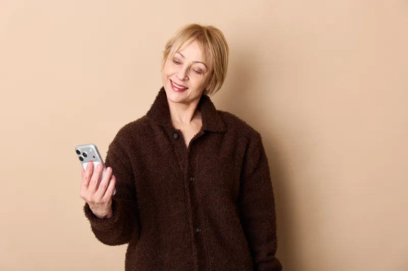 Smiling woman with short blonde hair holding a smartphone, studio portrait on beige background, showing communication and technology, relaxed casual lifestyle