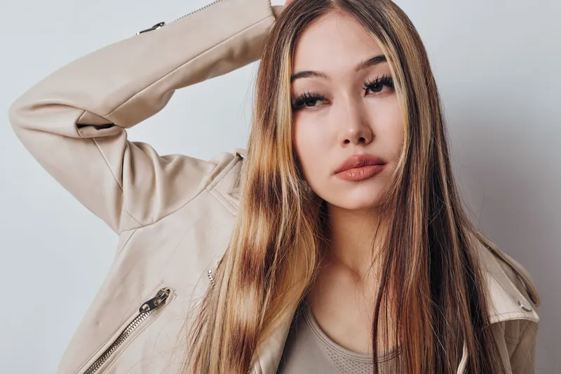 Young woman with long hair wearing beige jacket posing with hand behind head against white background. Portrait, fashion style and confident look with casual outfit and natural makeup.