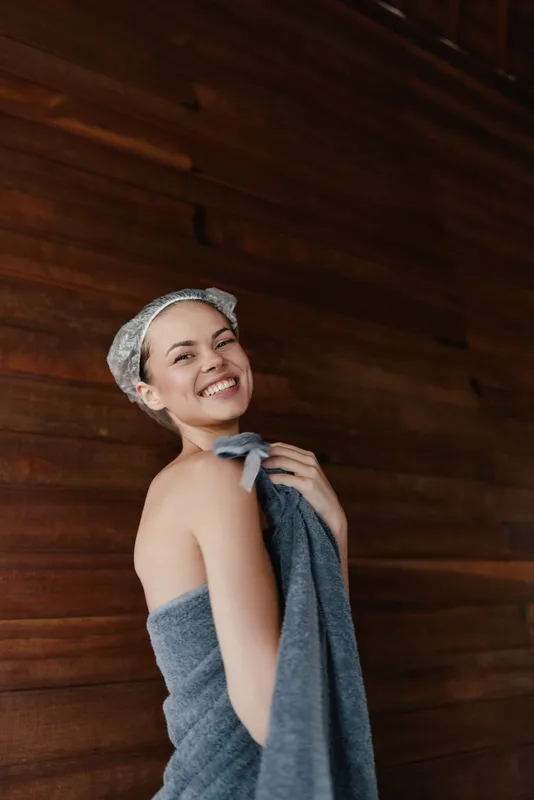 Smiling woman wrapped in towel with shower cap stands against dark wooden wall, expressing joy and relaxation after bath, natural skin, cozy atmosphere.