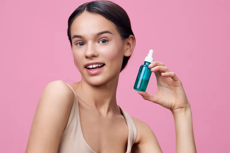 Young woman presenting a skincare serum on a pink background, showcasing beauty and confidence with a bright, fresh expression, ideal for wellness and beauty concepts