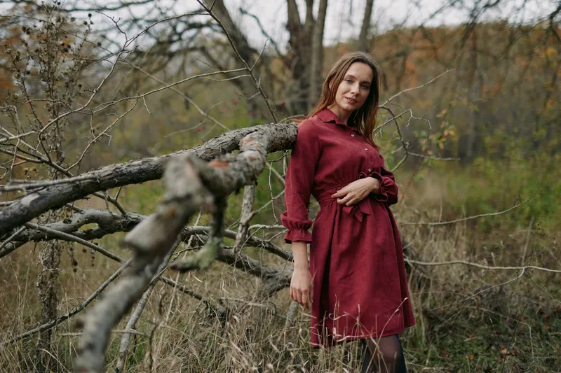 An authentic portrait of a woman in a deep red dress standing beside a weathered wooden fence in a natural field, conveying honest mood and quiet confidence.